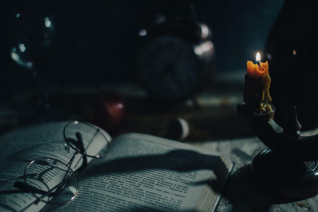 Dark image of open book with a candle in the upper right corner and a set of old-fashioned glasses in the middle of the book